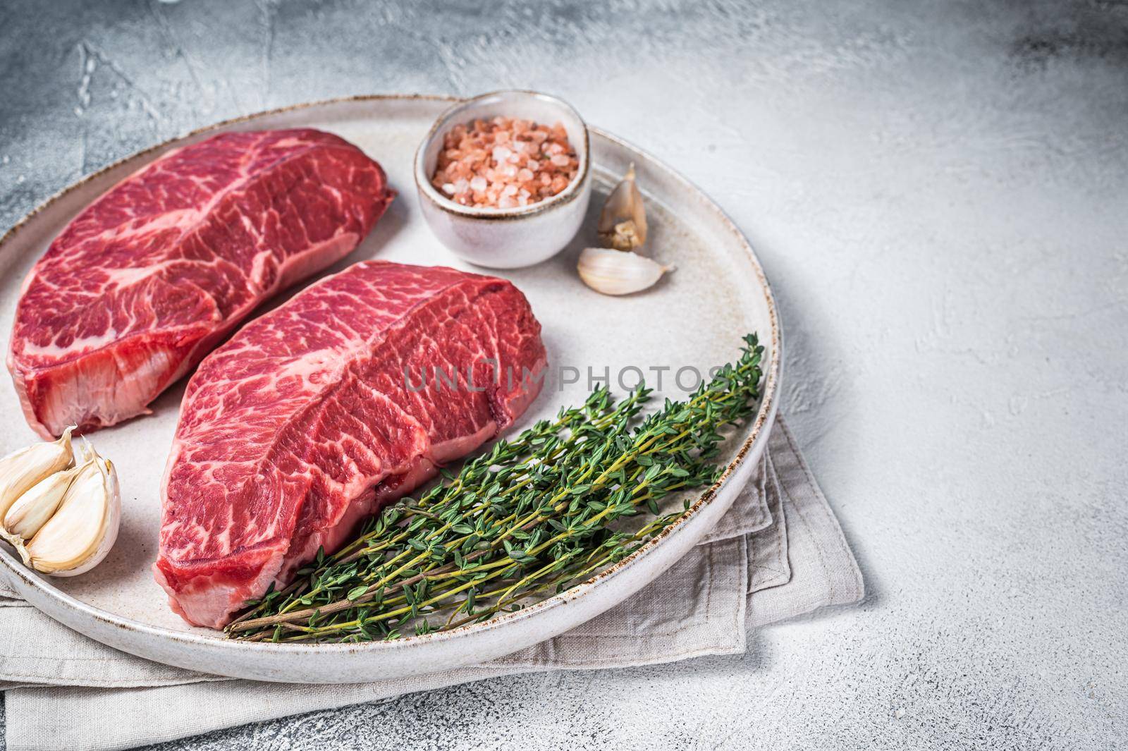 Raw Oyster Top Blade or flat iron roast beef meat steaks on a plate with herbs. White background. Top View. Copy space.