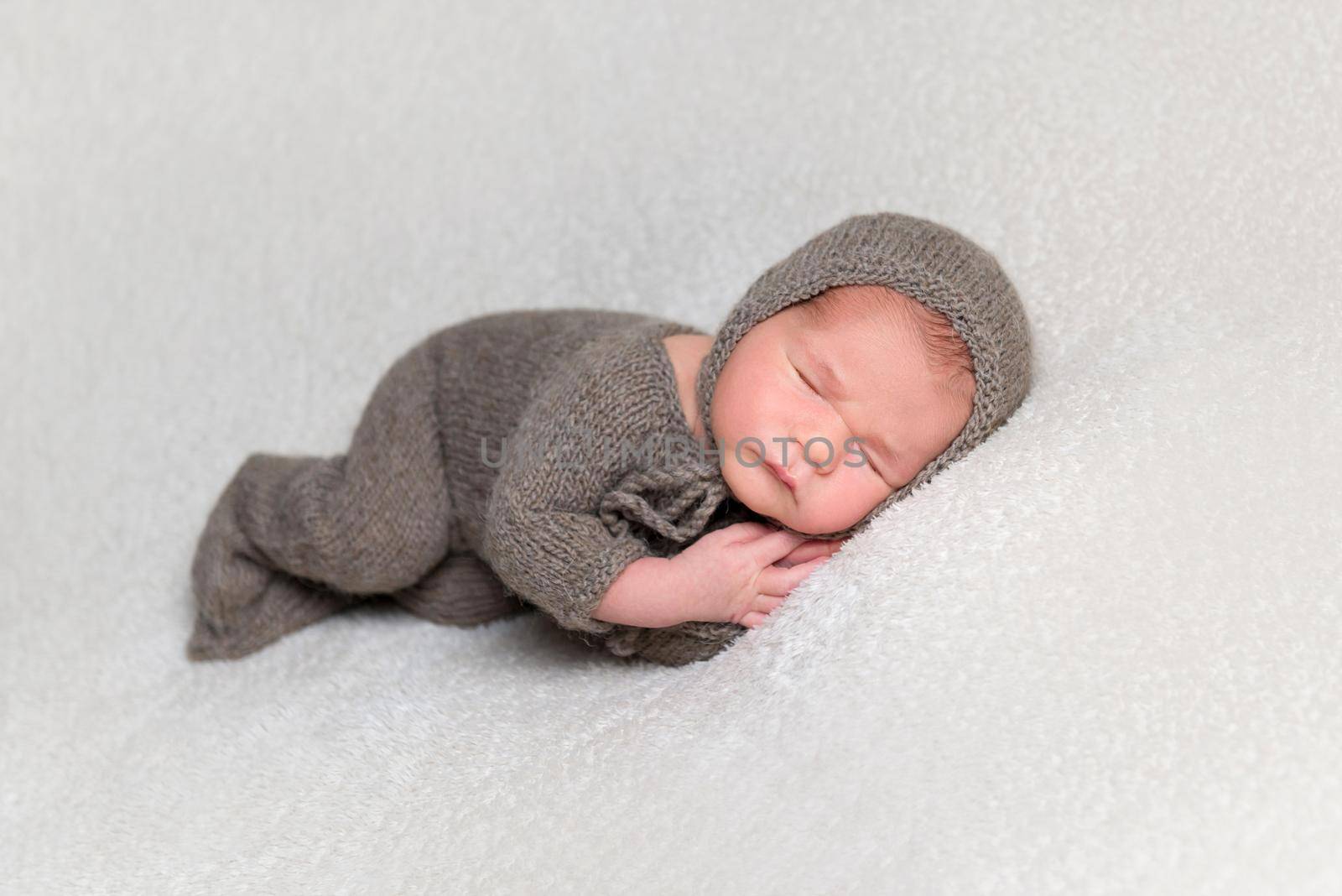 Dreamy baby boy wearing brown knitted clothes sleeps on his side on white background.