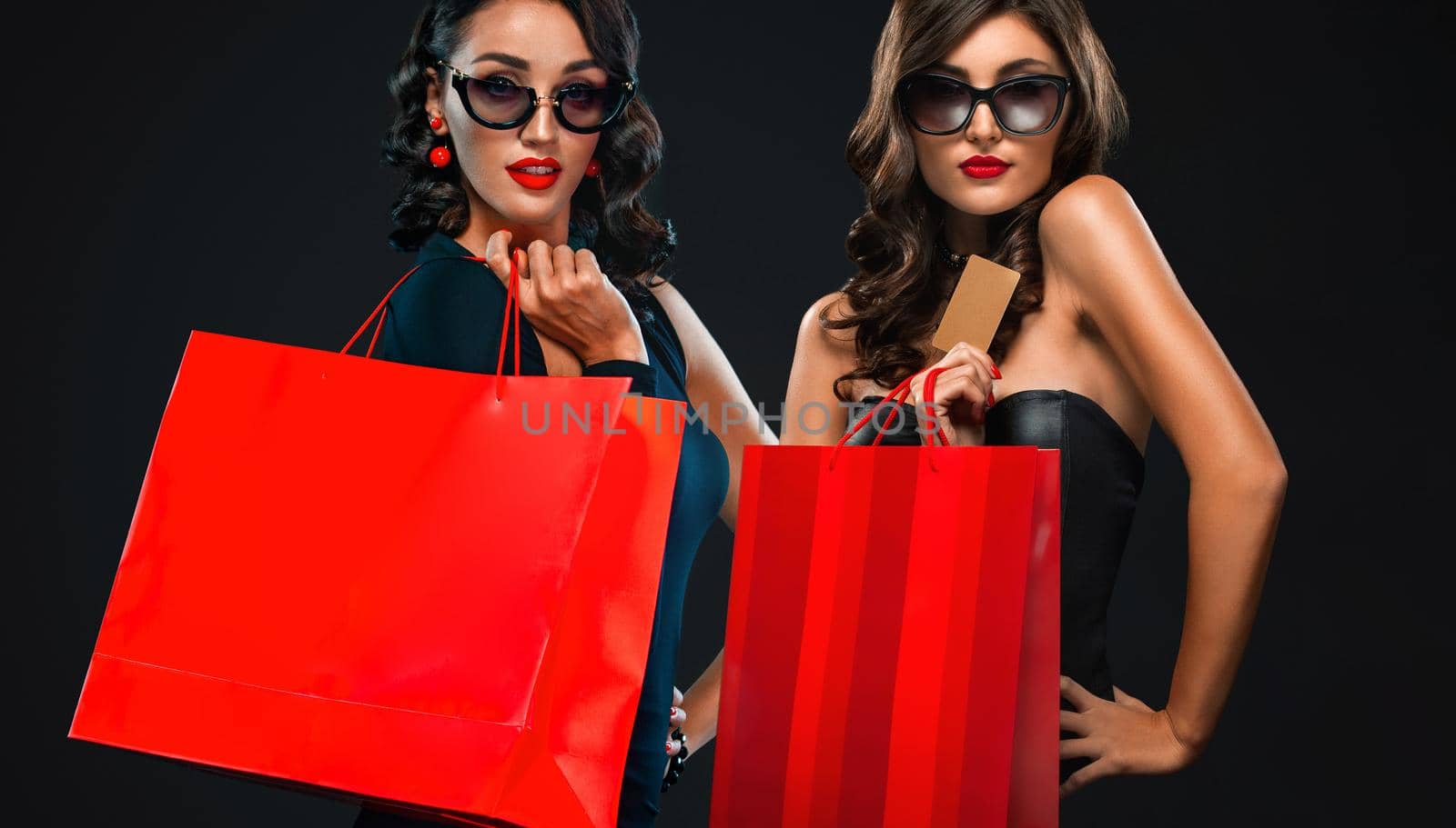 Black Friday sale concept for shops. Two woman in sunglasses holding red bag isolated on dark background at shopping mall. by MikeOrlov