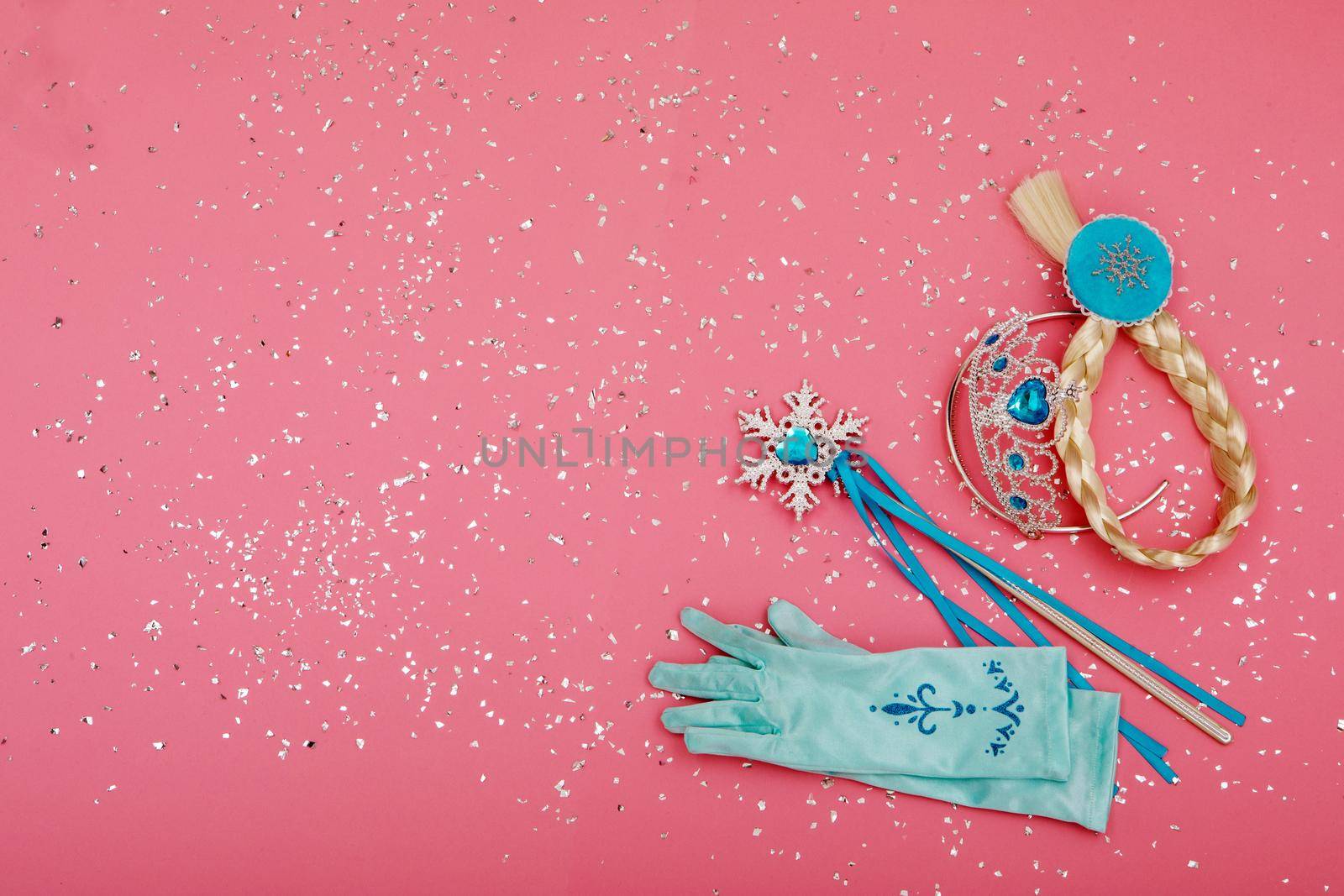 From above of magic wand and gloves placed near crown and pigtail on pink background with silver glitter