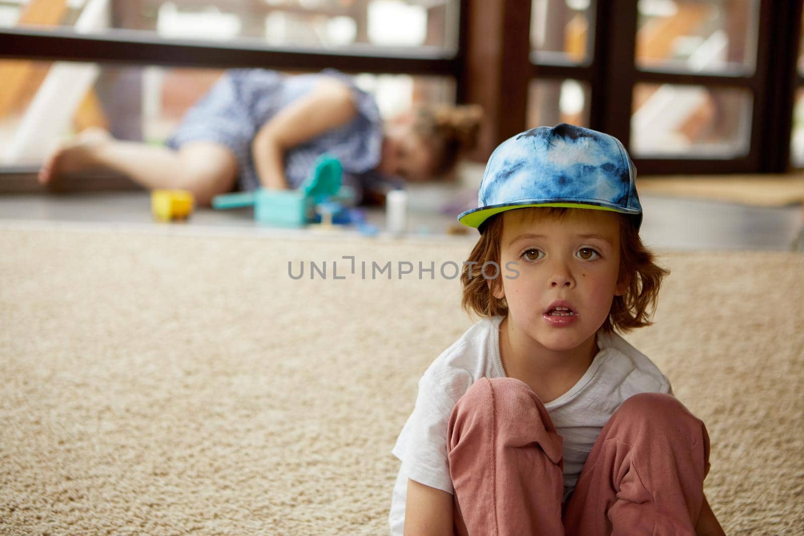 Adorable boy in casual clothes and trendy cap embracing knees and looking at camera while sitting on carpet in living room
