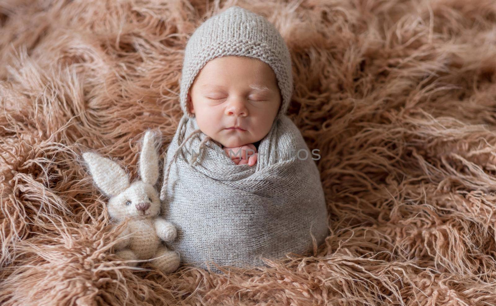 Little cute baby weared in gray knitted beanie and covered with gray knitted blanket sweetly sleeping with bunny nearby on the soft light brown bedcover