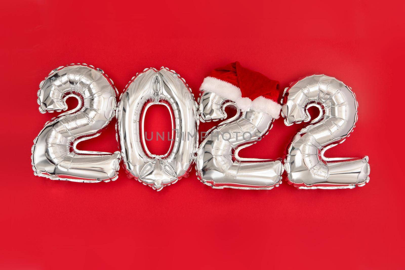 2022 inflatable balloons on red background with santa hat. Happy New year 2022 celebration. Silver foil balloons numeral 2022. Postcards and posters Copy space