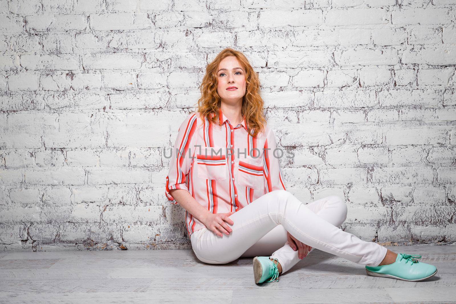 Young beautiful caucasian woman student is sitting on the floor near a white brick wall. Girl with red long curly hair with freckles on her face in bright summer clothes by Tomashevska