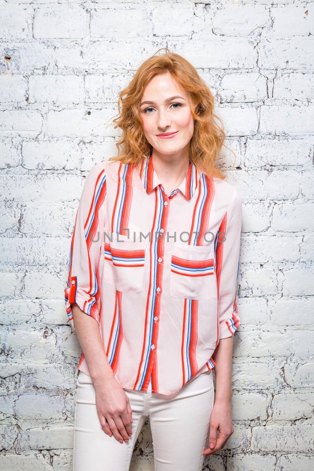 A beautiful young woman student with red curly hair and freckles is leaning against a brick wall of gray color. Dressed in a red striped shirt and white pants.