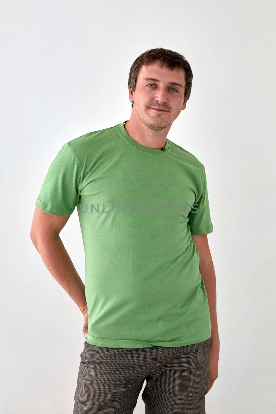 Positive young male in jeans with hand behind and green t shirt standing against white background and looking at camera