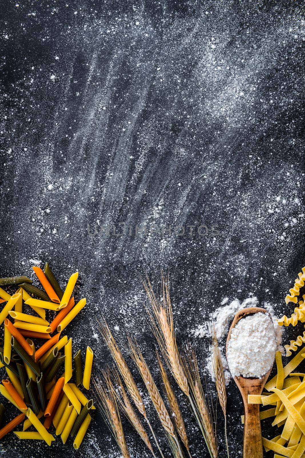 spikelets of wheat, pasta and spoon with flour on a black textured table