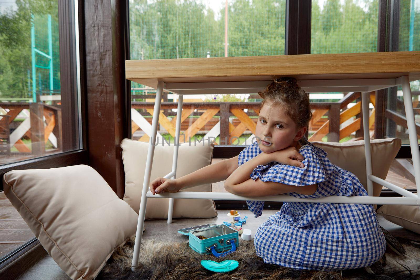 Girl in checkered dress looking at camera while sitting on floor near pillows and playing under table against large windows on weekend day