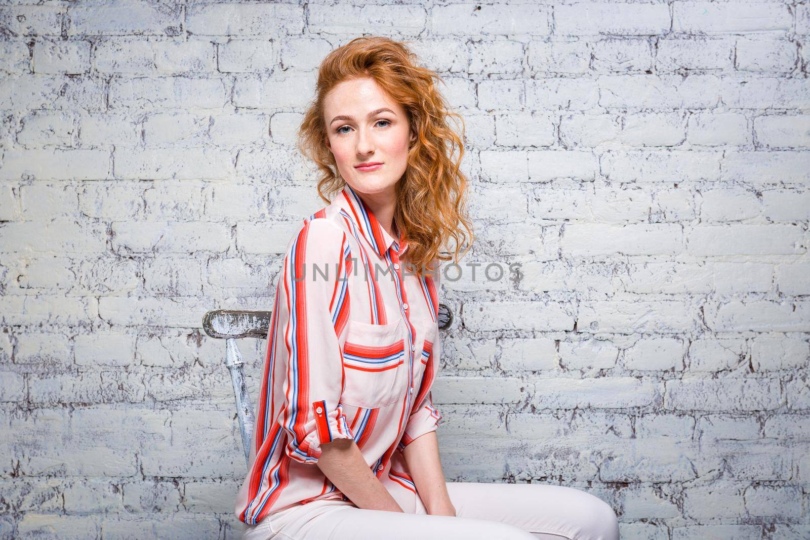portrait Beautiful young woman student with red curly hair and freckles on her face sitting on a wooden chair on a brick wall background in gray. Dressed in a red striped shirt.