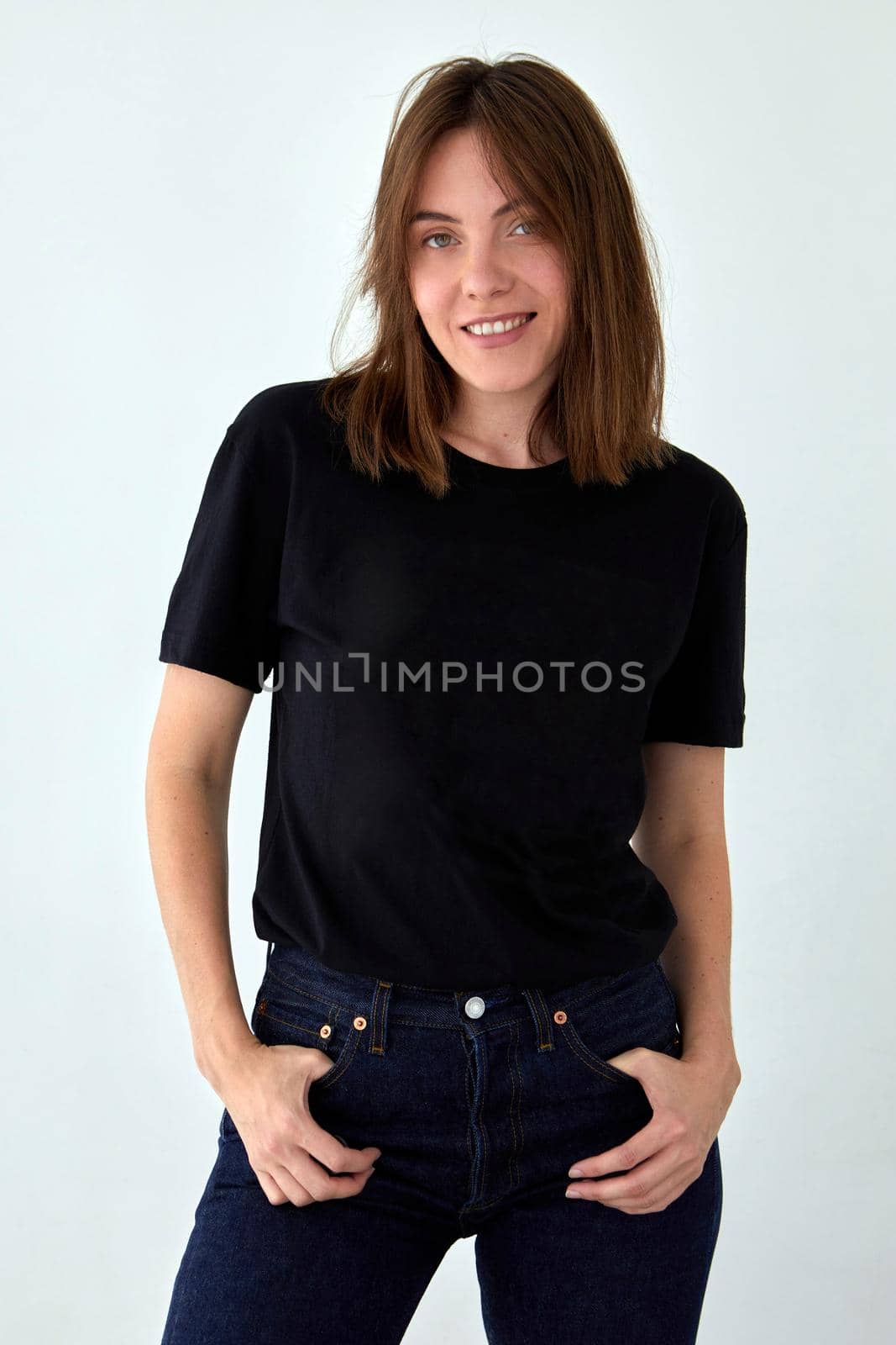 Positive female model wearing black t shirt and jeans standing with hands on waist against white background and looking at camera