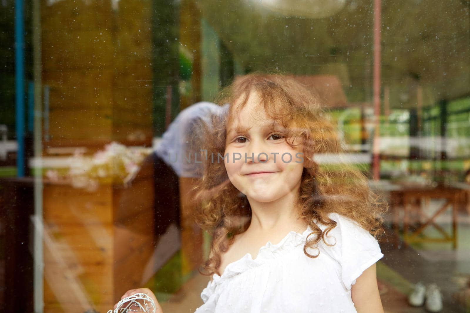Cute happy girl with curly hair smiling and looking at camera while standing behind transparent glass