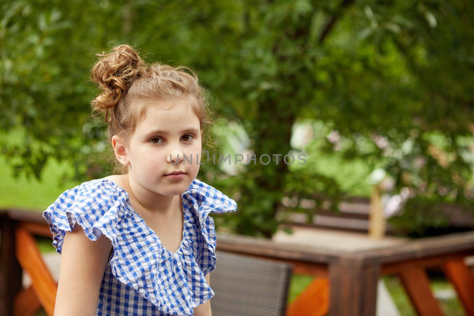 Adorable girl in checkered garment looking at camera while relaxing on patio on summer weekend day