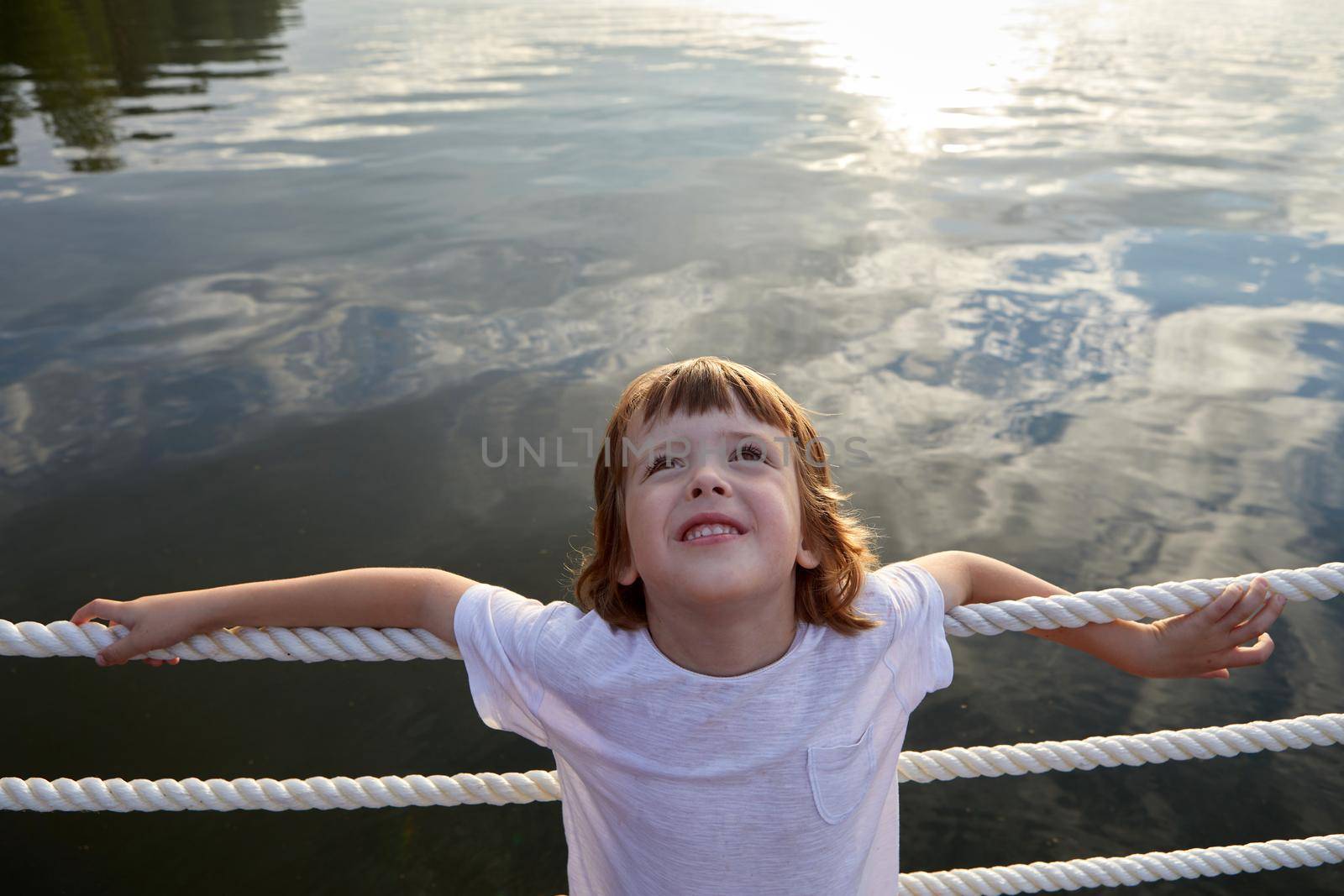 High angle of cute boy in white t shirt leaning on rope barrier and looking up while resting near clean pond water in summer