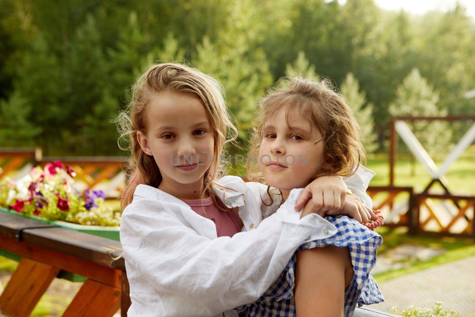 Adorable girl in white shirt embracing friend in checkered garment and looking at camera while resting near terrace railing on summer weekend day