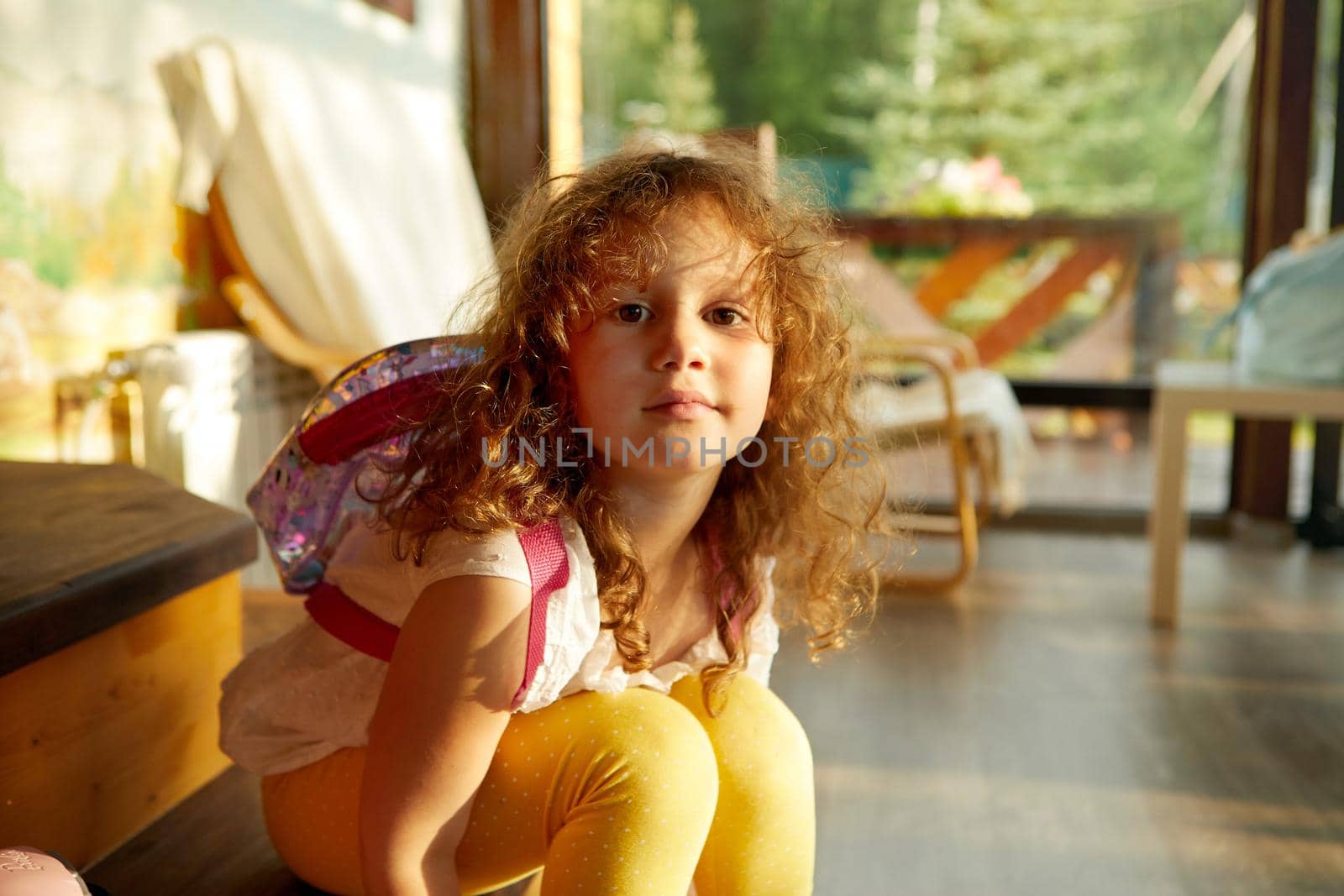 Adorable girl with backpack bending forward and looking at camera while sitting on steps in light room on weekend day. Child with a backpack going to school
