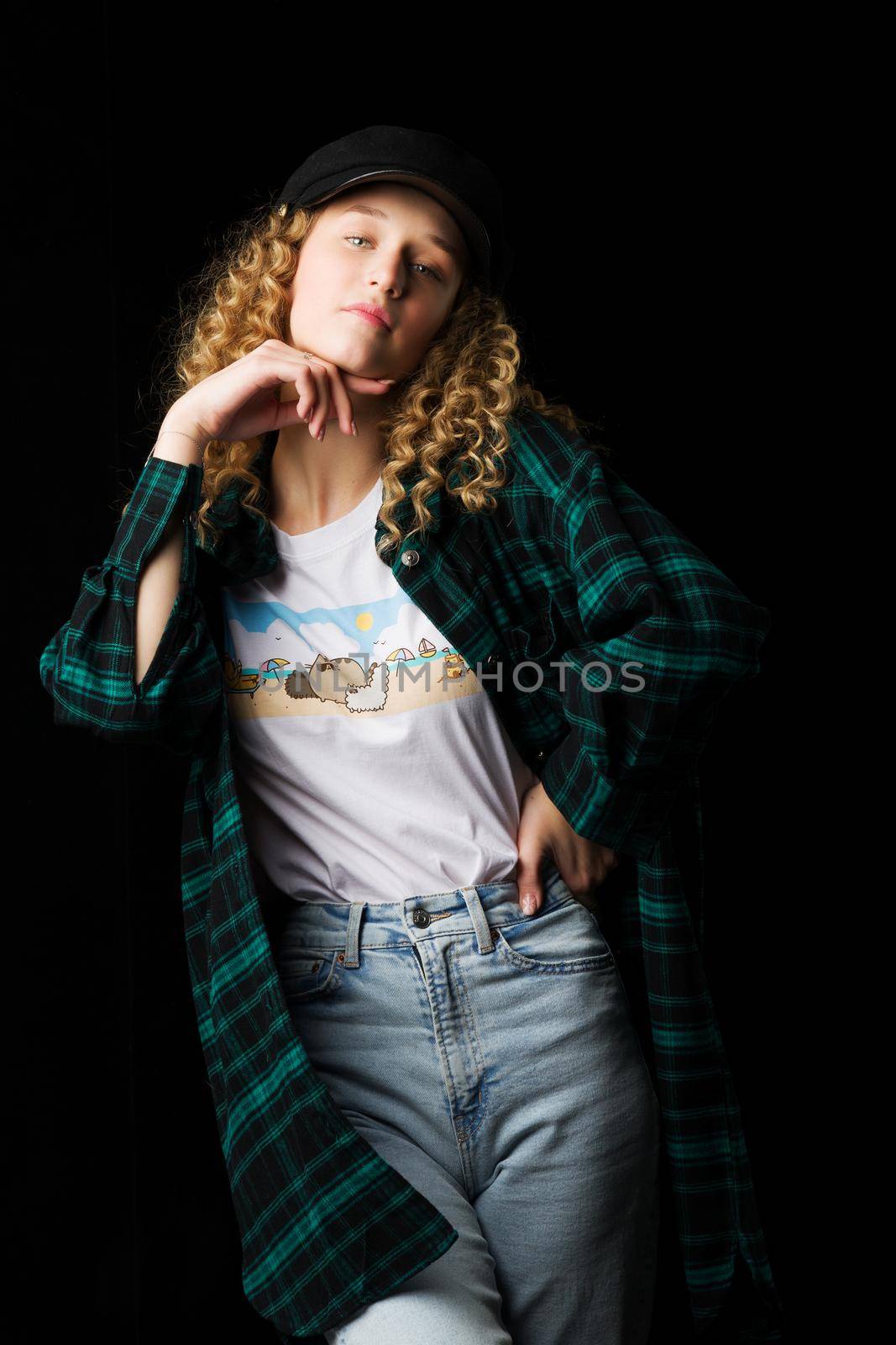 Charming girl posing keeping hand under her chin. Shot of stylish blonde curly girl in fashionable outfit looking at camera. Young woman wearing plaid shirt standing on black background