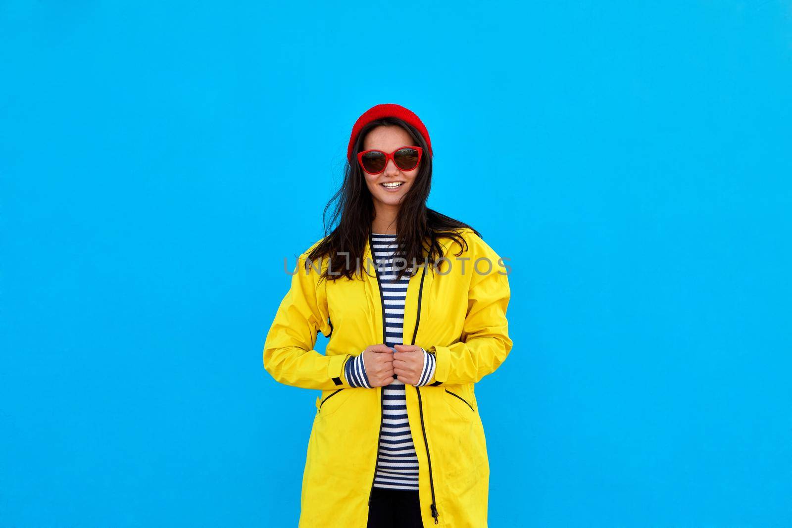 Smiling female traveler gesturewears hat, striped sweater and yellow raincoat, enjoys exciting travel, poses against blue background, copy space for text
