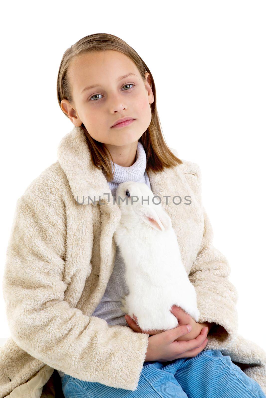 Stylish girl holding white rabbit. Pretty girl in beige fur coat hugging bunny pet animal. Close portrait of preteen child posing in studio against white background