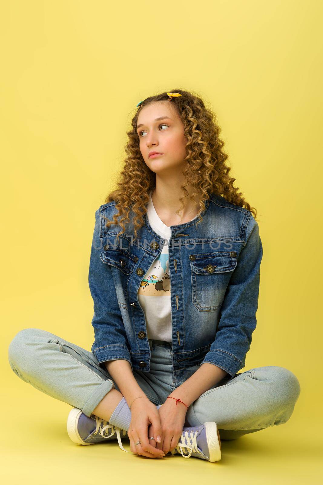 Pretty girl sitting on floor with crossed legs. Beautiful happy blonde girl wearing casual clothes standing with hand on her head against light yellow background.