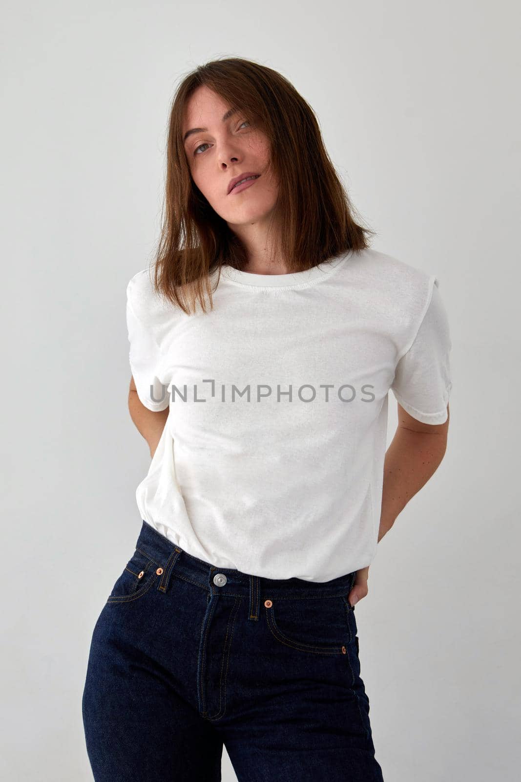 Positive female model wearing white t shirt and jeans standing with hands on waist against white background and looking at camera