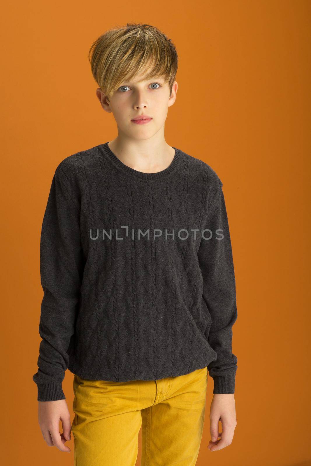 Stylish blond handsome teenage boy. Attractive boy wearing gray pullover and pants posing against ochre background. Portrait of stylish cheerful student teenager