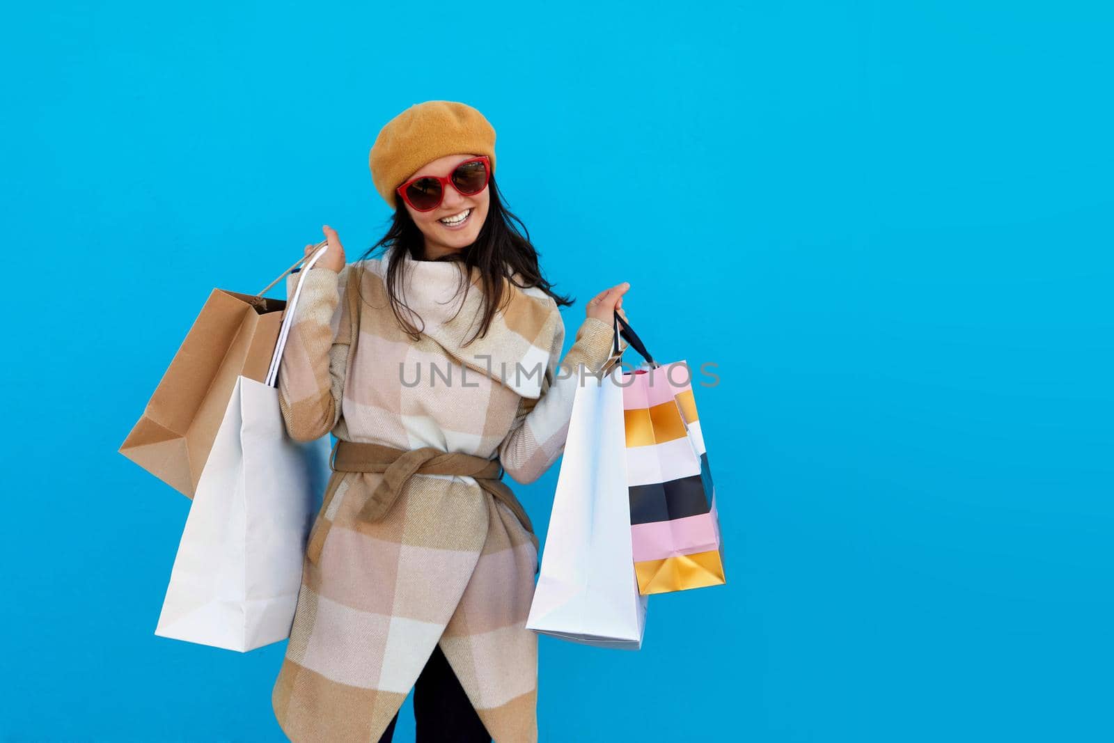 Cute attractive happy smiling stylish woman in sunglasses shopaholic in trendy beige coat and beret holding shopping bags blue studio background isolated colorful sale