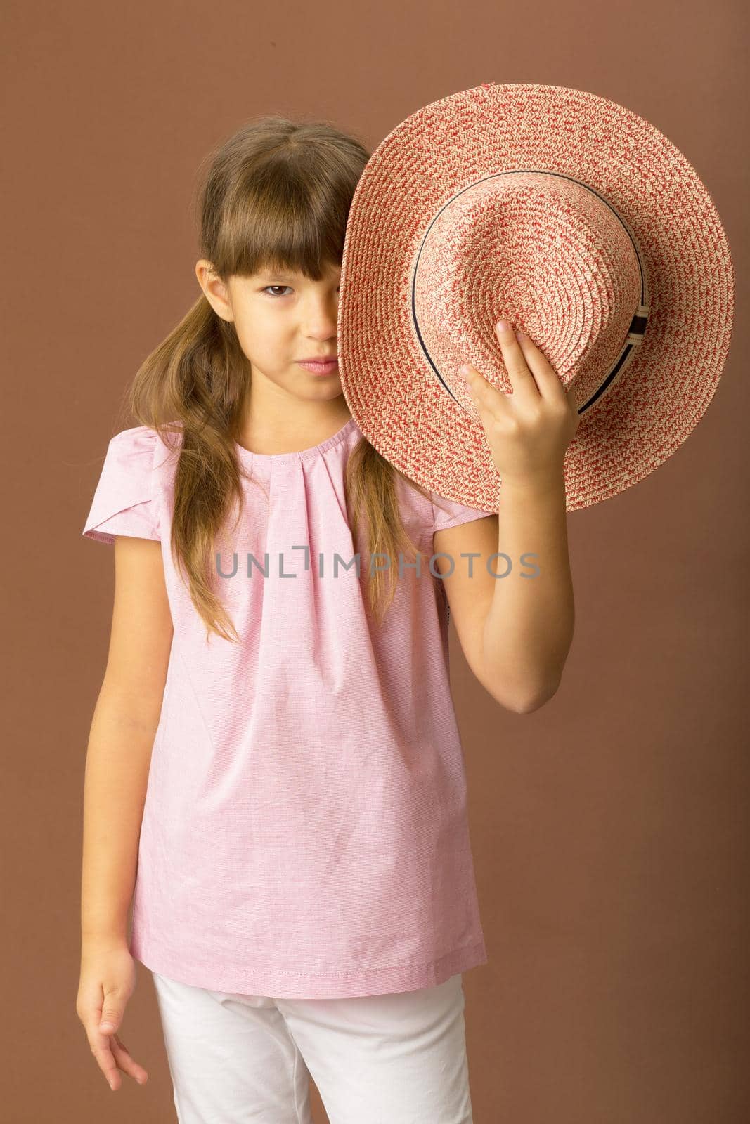 Girl in stylish summer clothes. A girl with ponytails in a pink blouse, white trousers covers her face with a straw hat. Portrait of a serious young child