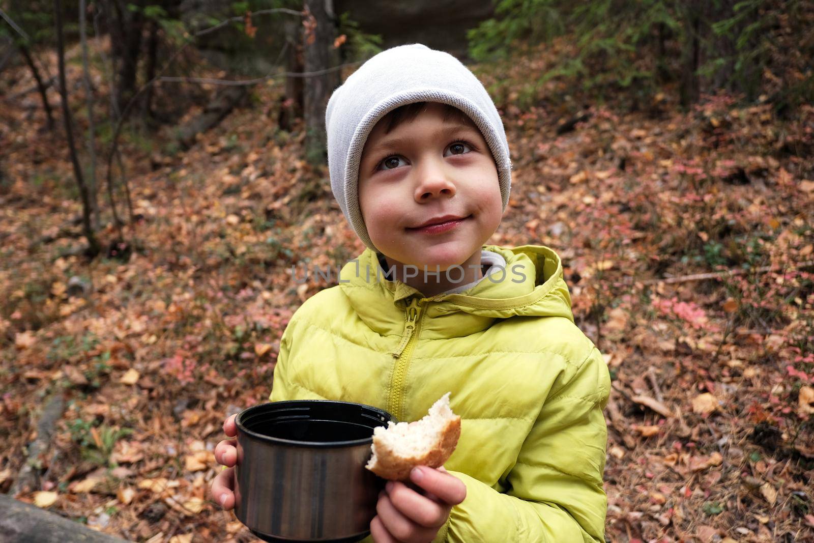 Positive boy in outerwear and cup of hot beverage and bread in hand looking away while sitting in woods during camping