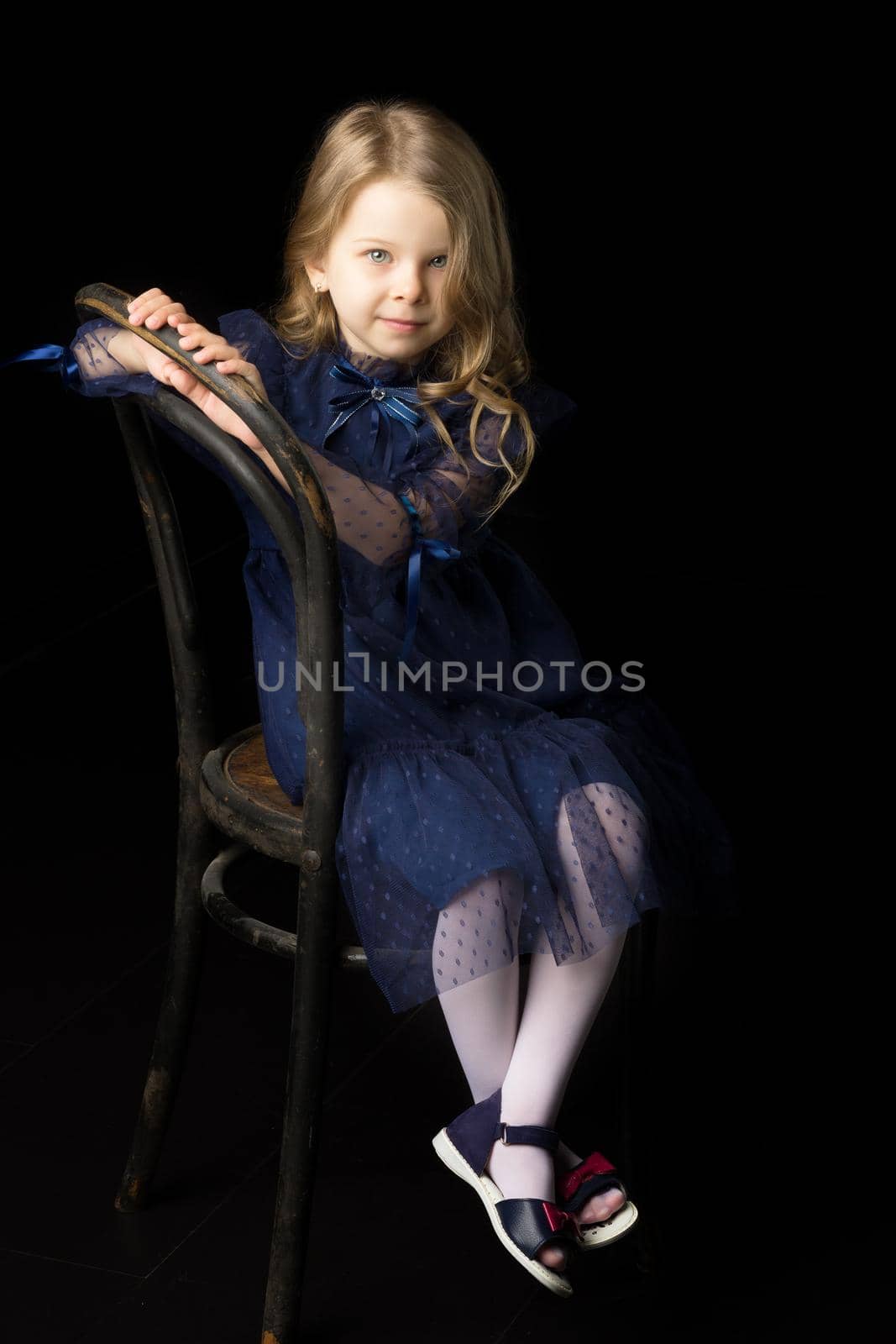 Close up portrait of happy sitting little girl. Lovely smiling blonde girl wearing fashionable clothes sitting on chair on isolated black background in studio.