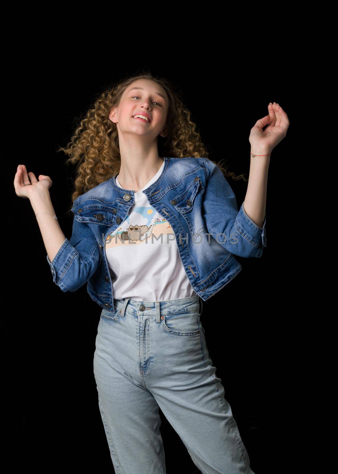 Happy joyful pretty girl in trendy denim outfit. Beautiful curly girl wearing jeans, denim jacket and white t-shirt laughing with her arms raised on isolated black background.