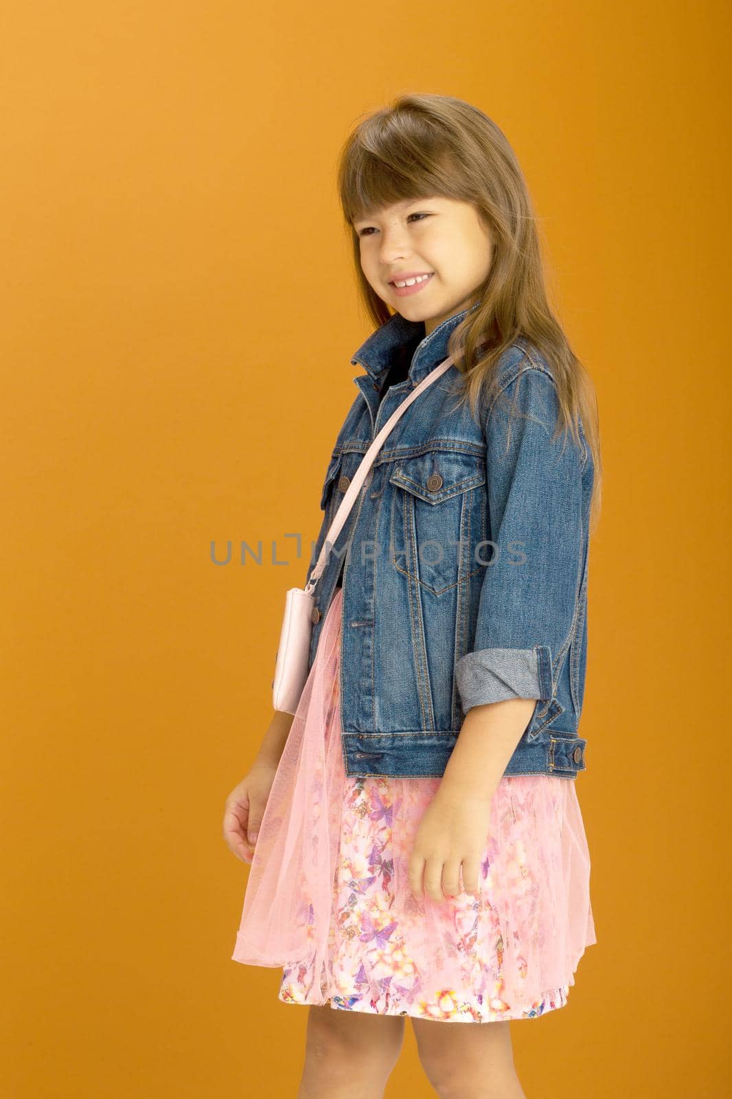 Beautiful girl in stylish outfit with shoulder bag. Smiling emotional preteen girl wearing denim jacket and pink fluffy skirt posing against ocher background with crossbody clutch decorated with heart