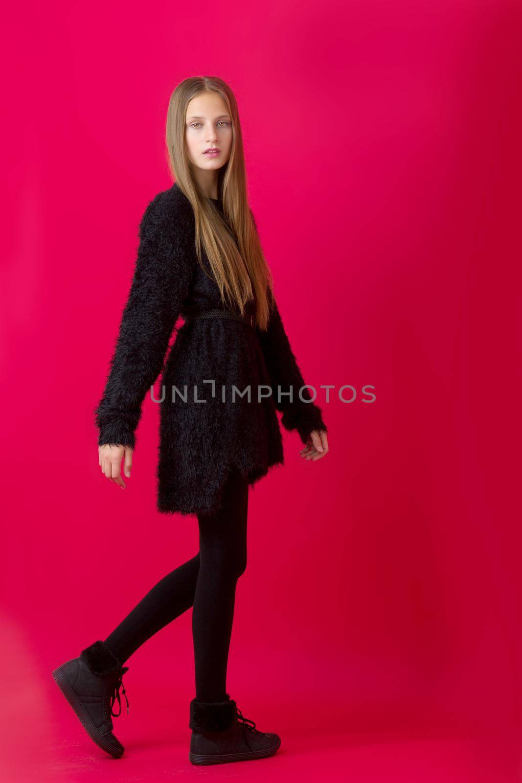 Beautiful stylish teenage girl in black outfit. Full length portrait of pretty girl with long brown straight hair wearing black dress, stockings and boots standing on red background in studio