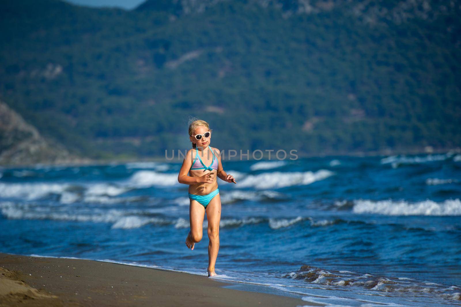 A little girl in a blue swimsuit and glasses runs on a Mediterranean beach in Turkey.