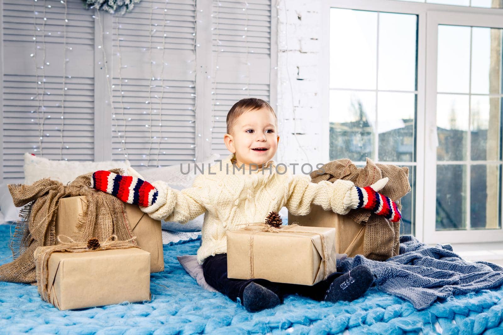 little funny playful boy a child sits on a bed on Christmas day with gift boxes in white wool knitted sweater and big bright mittens on it and laughs out loud. In interior there is a festive decor.