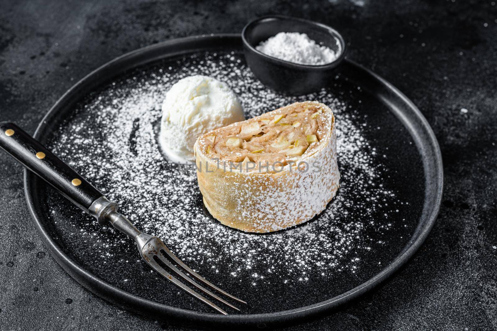 Apple strudel with cinnamon, powdered sugar and vanilla ice cream on a plate. Black background. Top view.