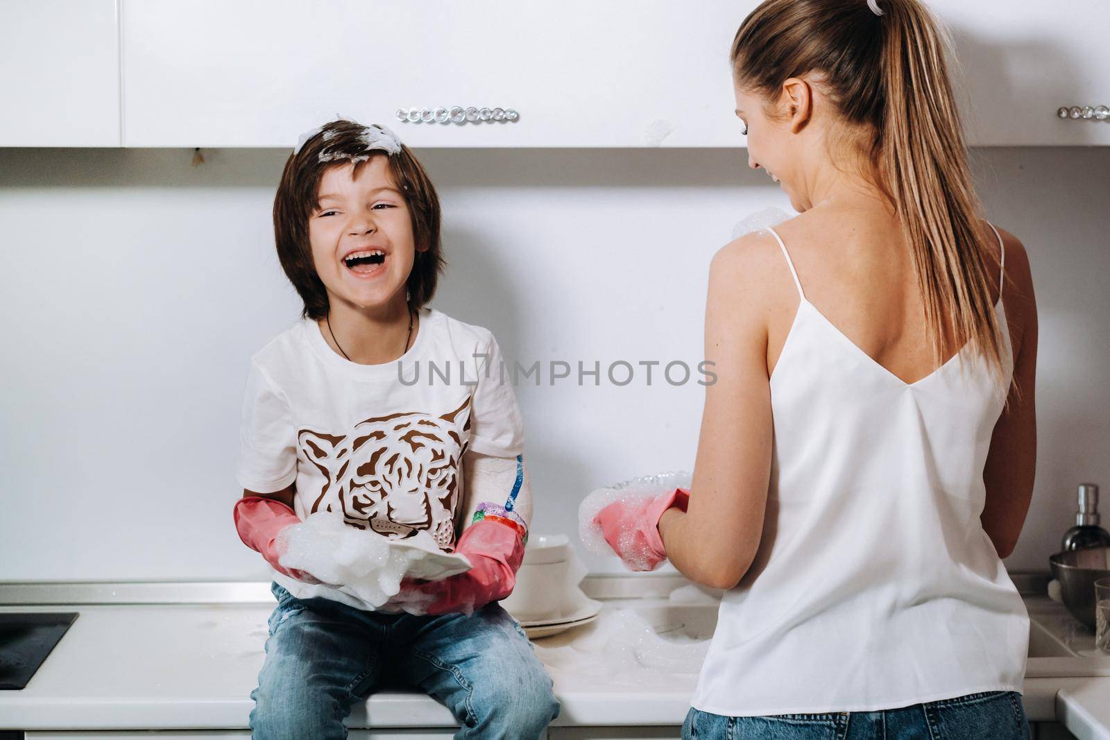 housewife mom in pink gloves washes dishes with her son by hand in the sink with detergent. A girl in white and a child with a cast cleans the house and washes dishes in homemade pink gloves.A child with a cast washes dishes and smiles by Lobachad