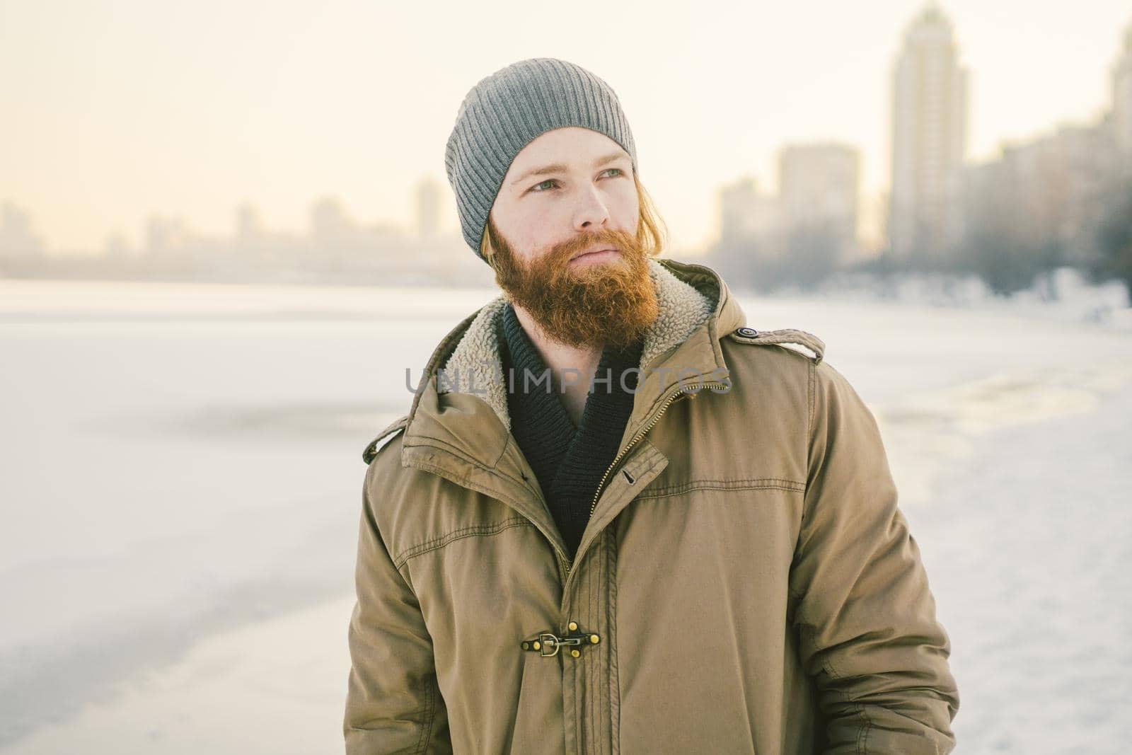 Close-up Caucasian young male red hair and beard in a hat and a park coat posing winter model against a background of a lake to freeze snow by Tomashevska