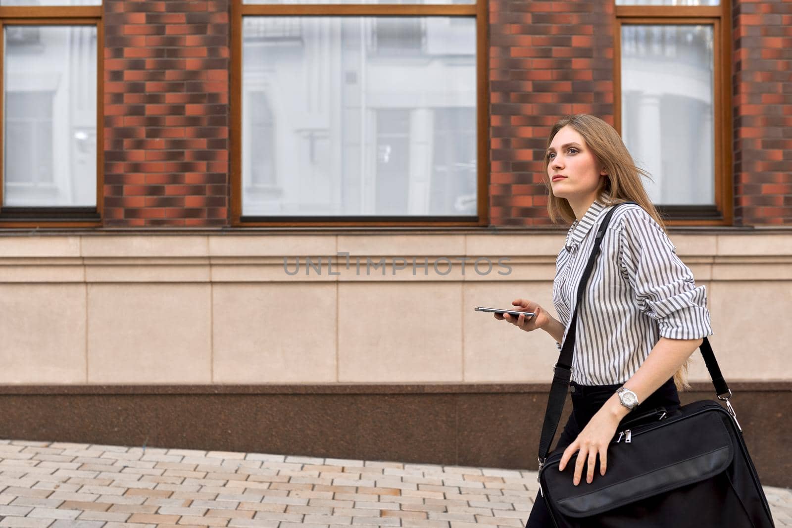 Young businesswoman walking talking on smartphone outdoor, street office building background, female professional walking forward profile view, copy space