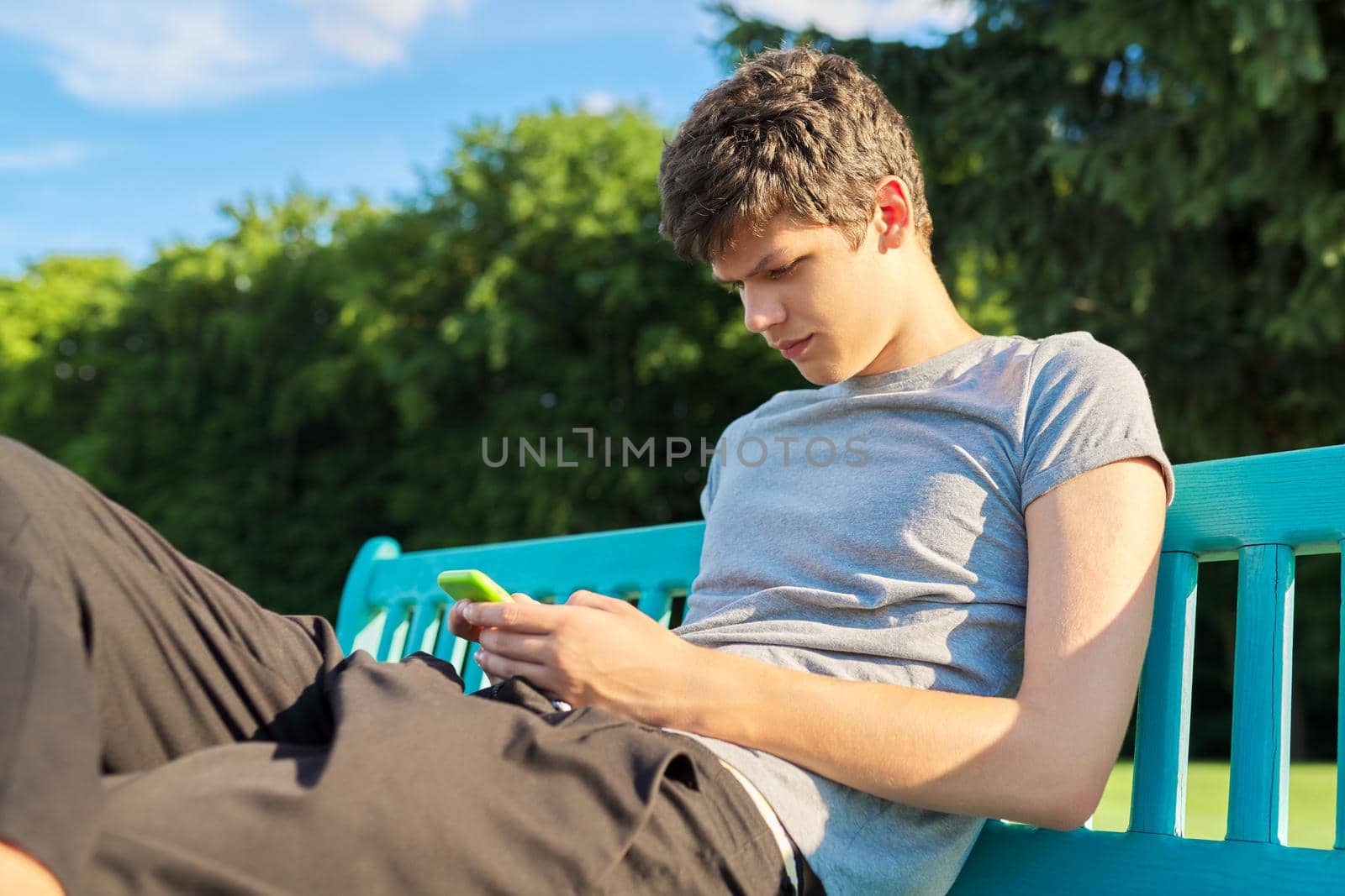 Serious guy teenager using smartphone, having rest, sitting on bench in park. Youth, technology, lifestyle, adolescence concept