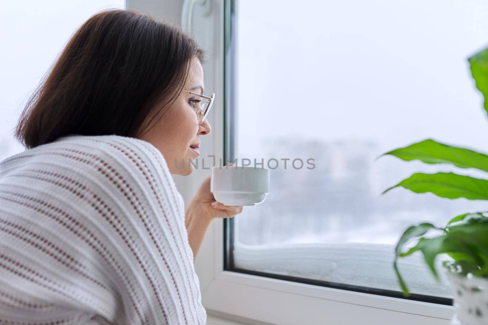 Winter season, snow, middle-aged woman looking out of home window at snowy winter city, copy space, christmas holidays, weather, nature, lifestyle concept