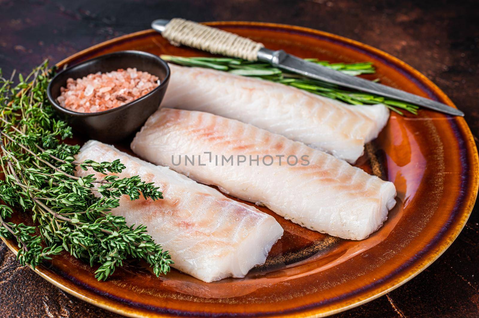 Raw cod loin fillet steaks on rustic plate with thyme and salt. Dark background. Top view.