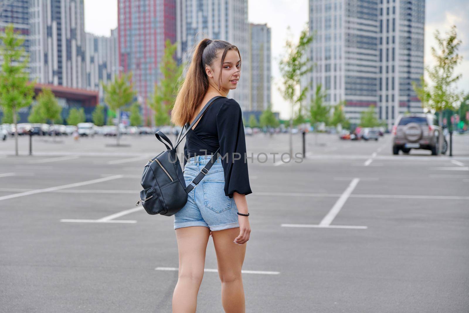 Fashionable beautiful smiling teenage girl in shorts with backpack looking at camera in big city parking lot, urban style, fashion, beauty, adolescence concept