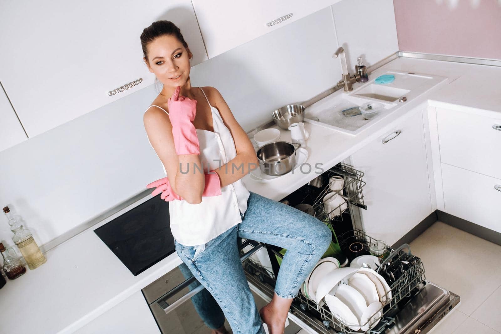 a housewife girl in pink gloves after cleaning the house sits tired in the kitchen.In the white kitchen, the girl has washed the dishes and is resting.Lots of washed dishes.