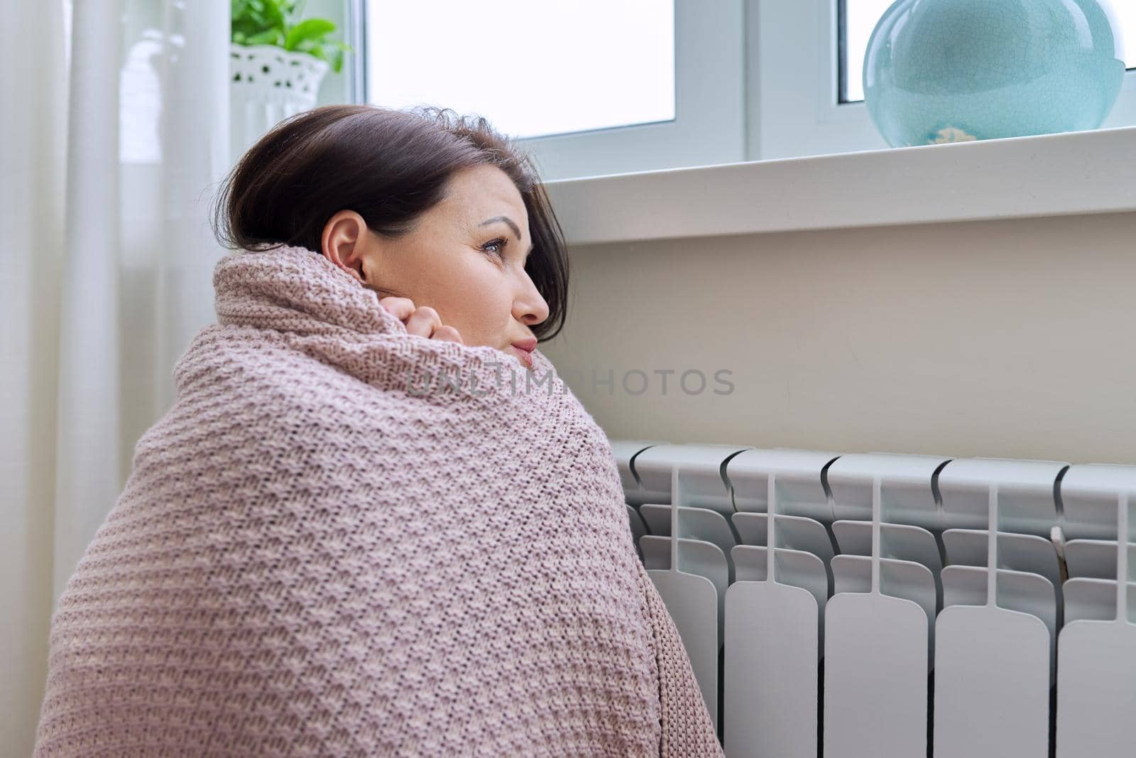 Winter, heating season. Woman in warm sweater sitting in home room near heating radiator by VH-studio