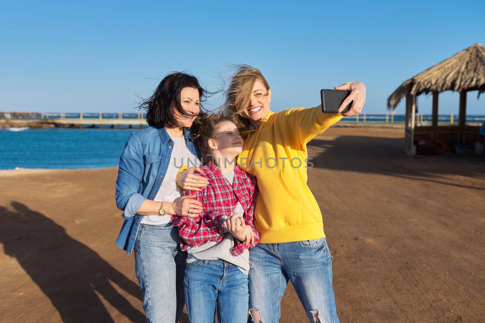 Happy two middle aged women and girl child taking selfie on smartphone, sea beach background by VH-studio