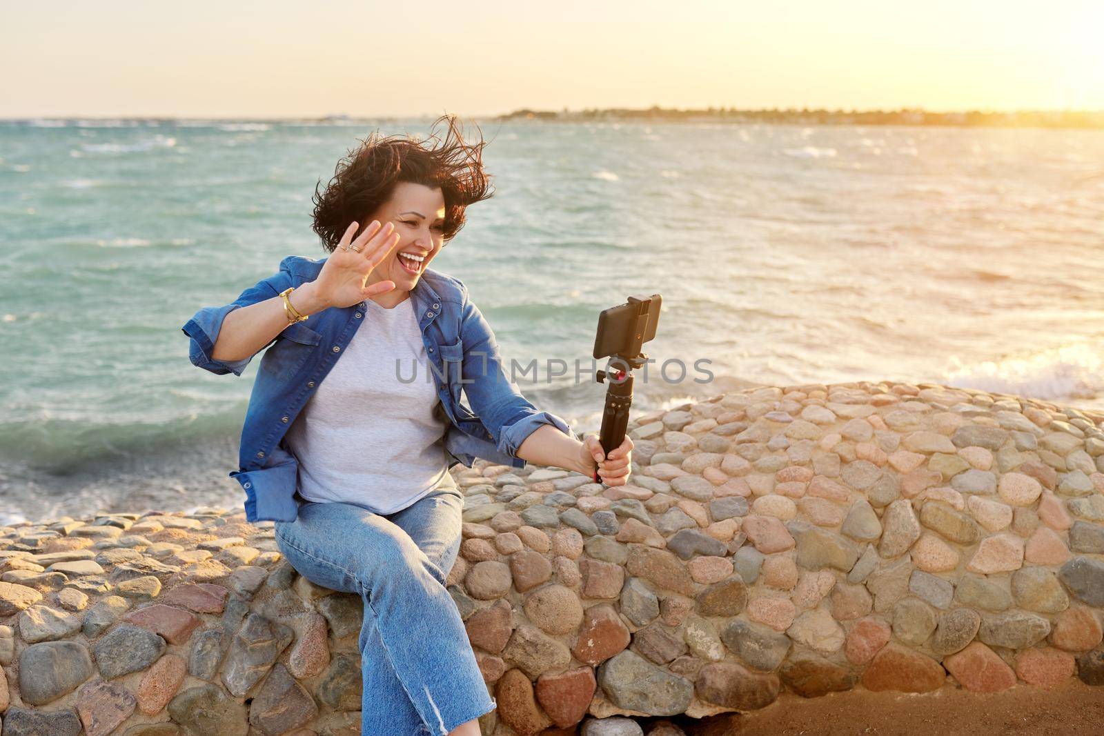 Middle-aged female making online video call talking laughing, sunset seascape background by VH-studio