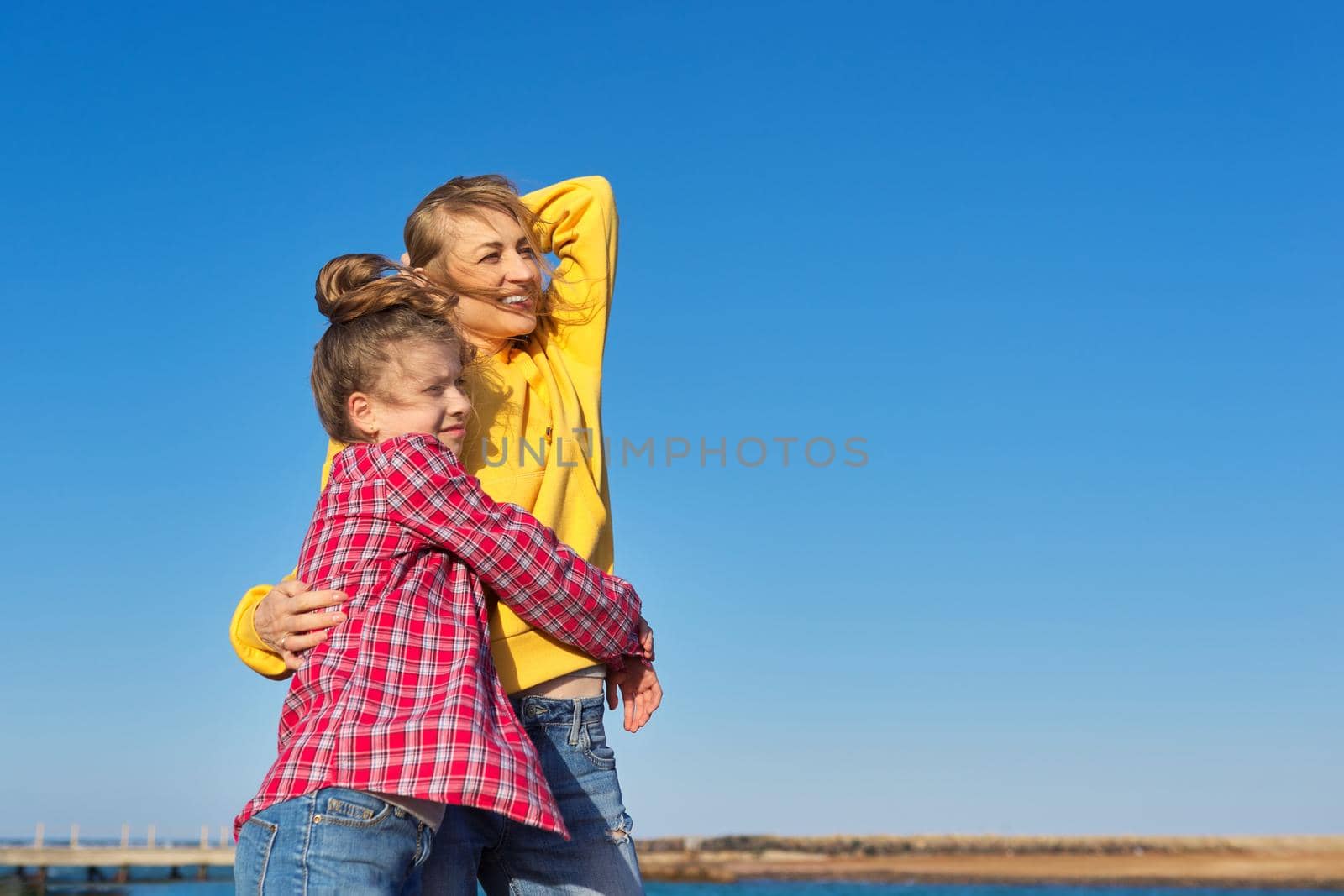 Happy mom and daughter child on seashore, relaxing on sandy beach, autumn winter spring season, copy space, blue sky background. Family vacation, travel, parent child relationship, happiness, joy