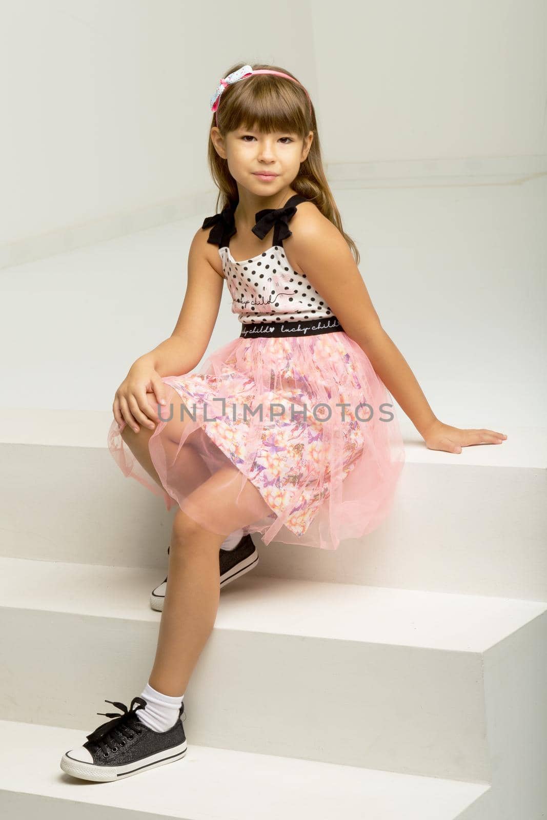 Stylish girl sitting on white staircase. Girl wearing nice dress and sneakers sitting on step leaning back on her hands and bowed her head. Beautiful child looking seriously at camera
