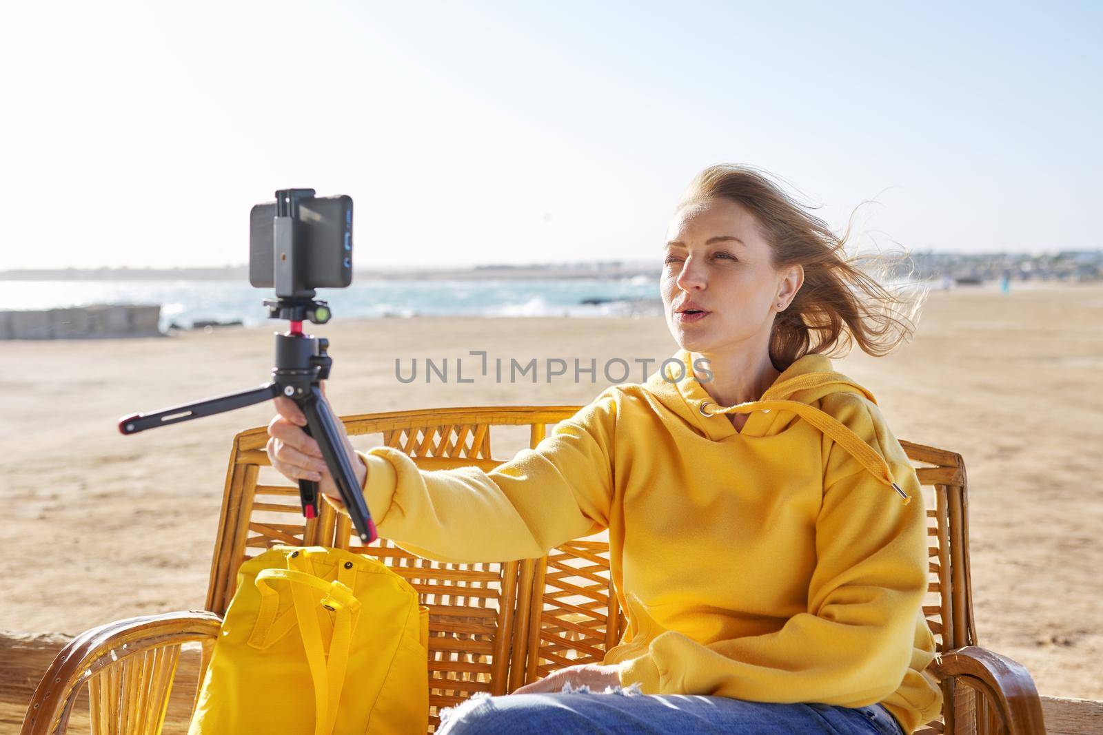 Technology, lifestyle, leisure and travel. Middle-aged female making online video call talking laughing, using smartphone, sunset seascape, sandy beach background, tropical autumn winter spring season