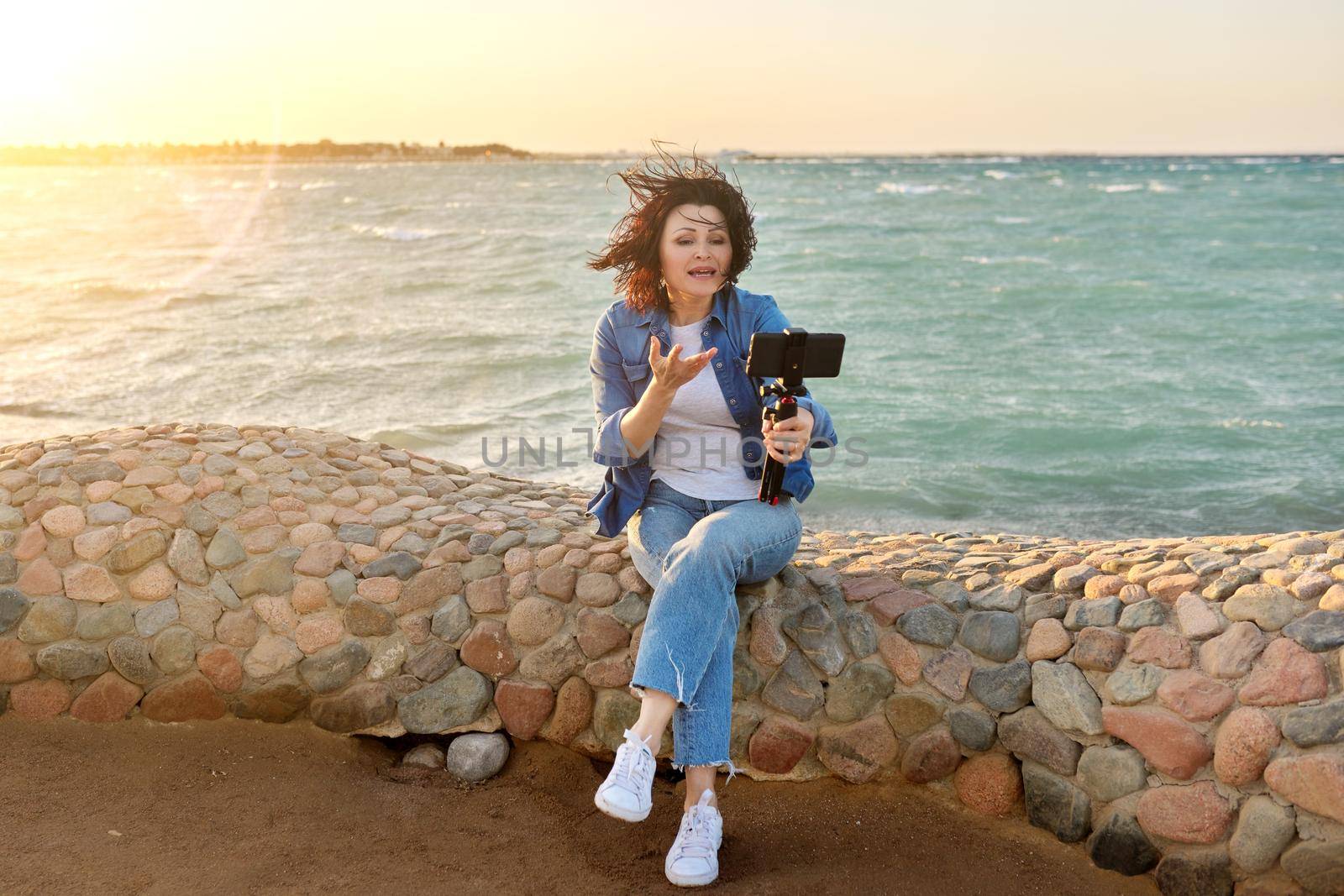 Technology, lifestyle, leisure and travel. Middle-aged female making online video call talking laughing, using smartphone, sunset seascape, sandy beach background, tropical autumn winter spring season